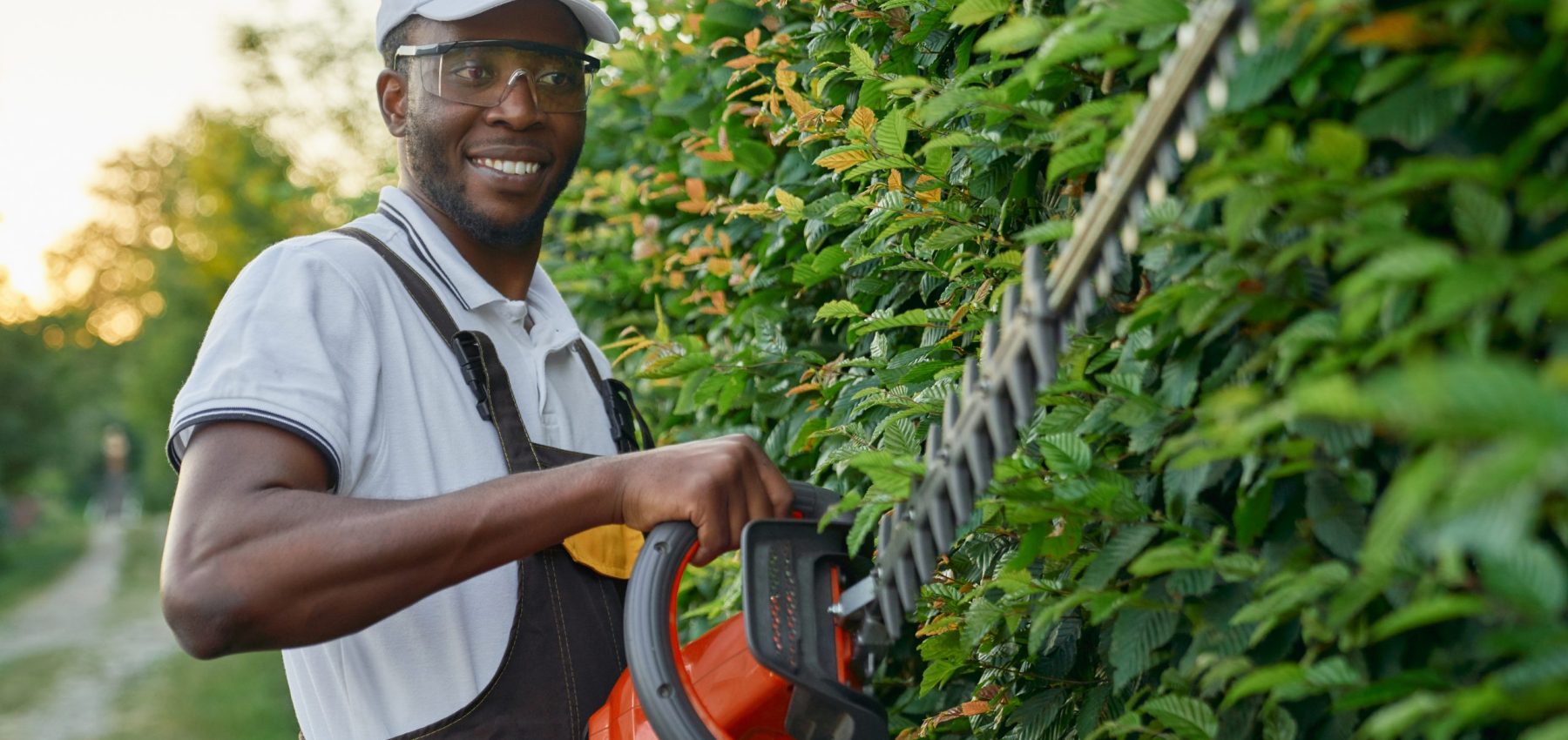 Smiling african gardener wearing special overall with protective glasses working with professional garden equipment outdoors. Young man using hedge trimmer at work